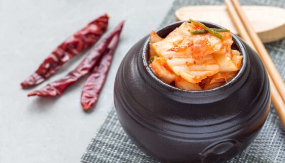 A pot of kimchi on a table with chopsticks and chilis